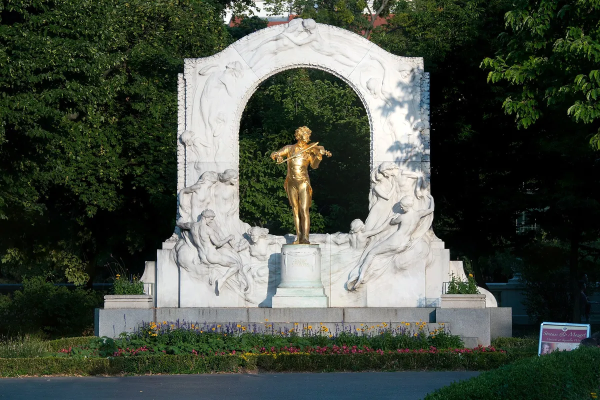 A gilded bronze figure of a violinist stands under a white marble arch with relief figures, in a green city park