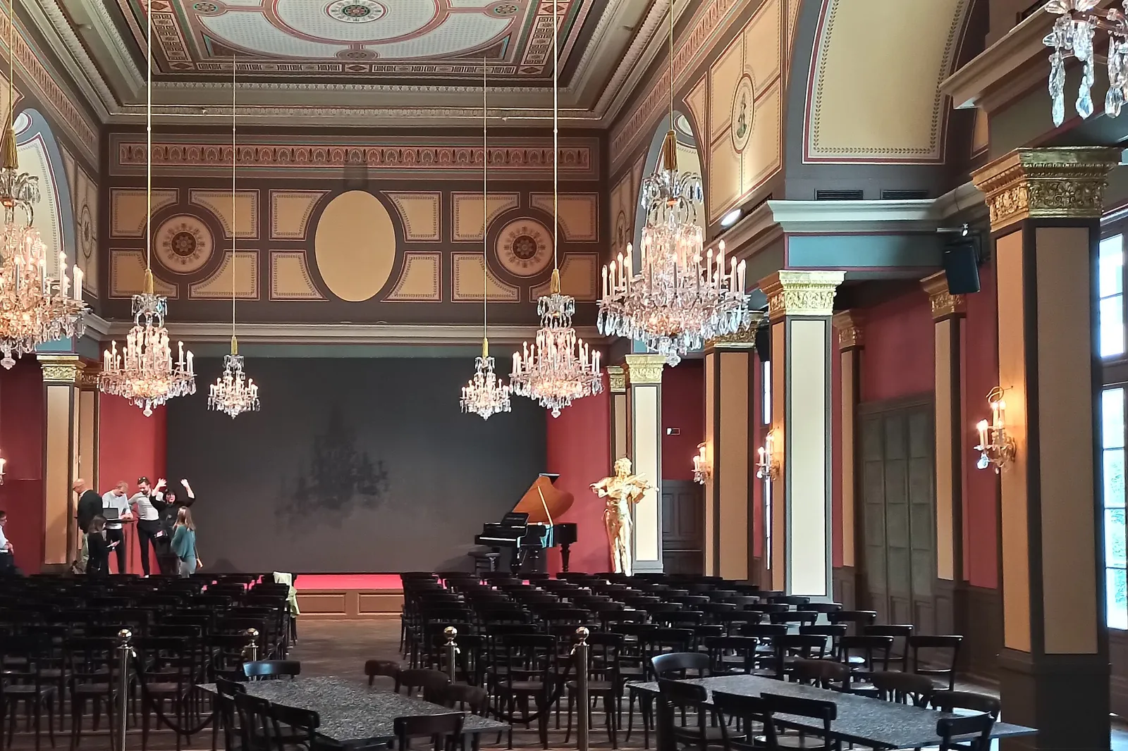 The stage end of the hall: a grand piano and a gilded violinist figure stand before a dark screen under crystal chandeliers